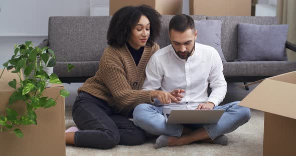 Concentrated Caucasian Man and Happy Afro American Woman Sitting on Living Room Floor Near Cardboard