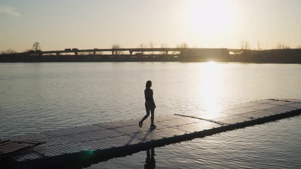 Young athletic girl goes down the pontoon along the lake on the background of beautiful sunset