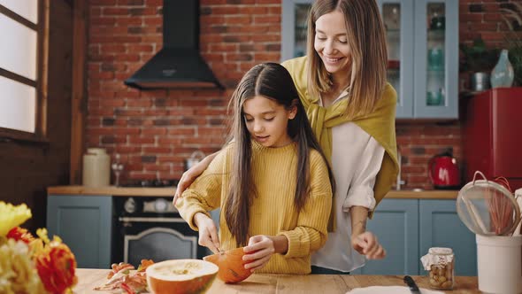 Little Girl Separating Pumpkin Seeds From Pulp with Spoon and Smiling Mom Helping Her