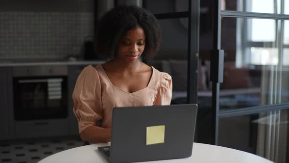 Smiling Young African Woman Sitting at Table in Cozy Light Warm Kitchen Working on Laptop Looking to