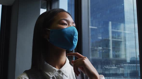 Thoughtful asian woman wearing face mask looking out of window at modern office