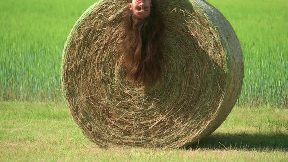 A beautiful woman playing and laying upside down on a large bale of hay on a farm in France.