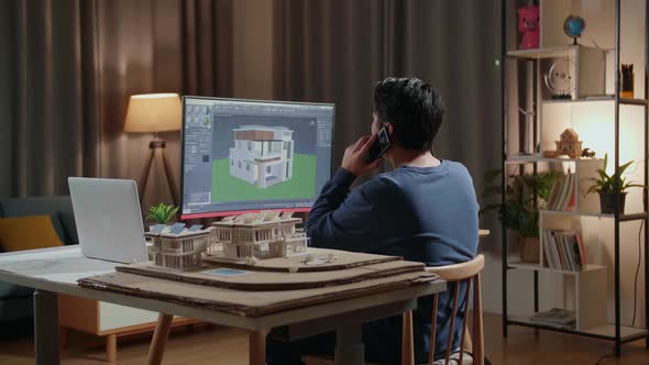 Asian Male Engineer Talking On Smartphone While Designing House On A Desktop At Home