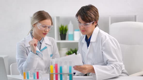 Two Female Chemist Wearing Uniform and Protective Glasses Looking on Paper Document in Lab