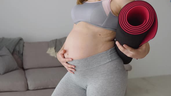 Pregnant Woman in Sportswear Standing with Fitness Mat in Hands at Home in Living Room