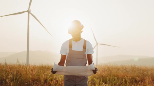 African Man with Blueprints in Hands Among Farm with Wind Turbines