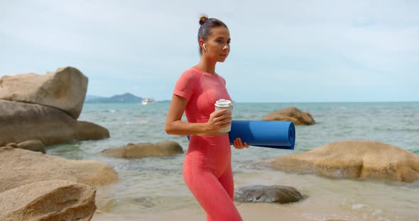 Young Woman Walks on the Beach for Fitness Exercise She Holds Yoga Mat and Coffee Cup in the Hand