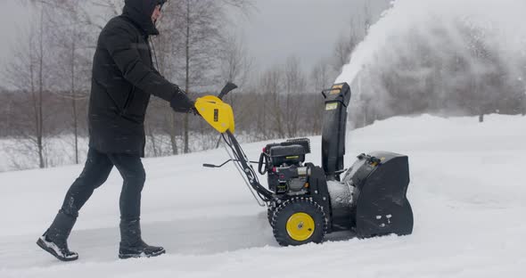 Man Cleans Snow With Snow Plow Background Of Wooden House After Heavy Snowfall