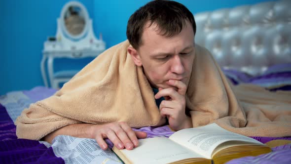 Attractive young man reading a book while lying on a bed in a bedroom