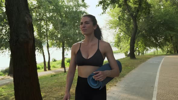 Young strong woman holding yoga mat in park outdoors