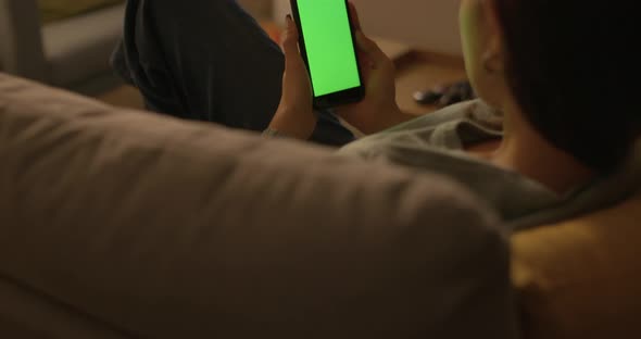 Young woman sitting on the couch at home and using her touch screen smartphone, green screen mock-up