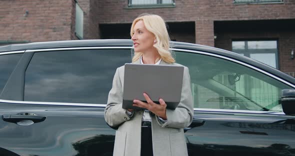 Attractive Blonde Female Driver Standing Near Her Car Using Laptop on a City Street.