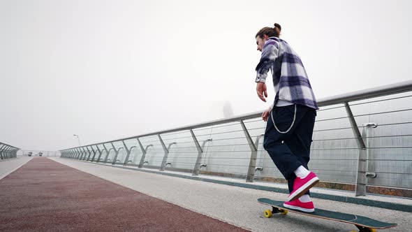 Fashionable Hipster in a Plaid Shirt and a Ponytail on His Head Rides a Skateboard on the City