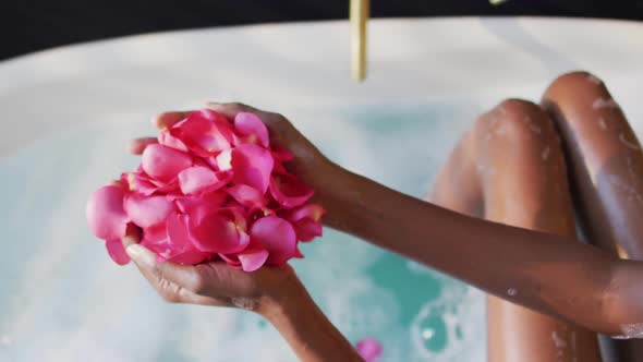 African american woman taking bath and touching flower petals in water in bathroom