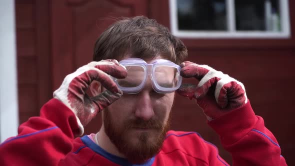 Worker Young Bearded Man in Red Working Gloves and Red Clothes Put on Protective Glasses and Look in