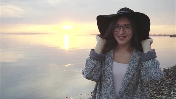 Young Pretty and Smiling Woman in Black Hat and Glasses Near the Sea at Sunset
