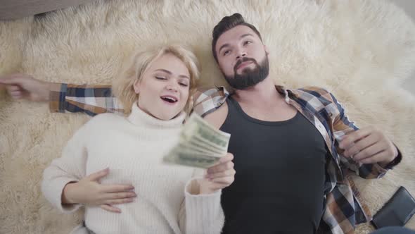 Blond Positive Caucasian Woman Taking Cash From Her Husband and Kissing Him on Cheek. Rich Spouses
