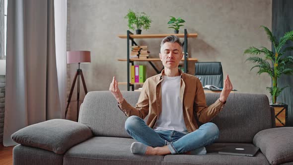 Peaceful Middle Aged Man Meditating at Home Sitting on Couch in Lotus Position and Practicing