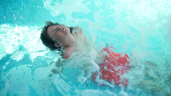 Top View of Playful Cute Boy in Blue Bubbling Water in Swimming Pool Smiling and Looking at Camera