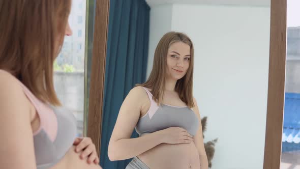 A Pregnant Woman Stands in Front of a Mirror and Holds Her Hands on Her Belly