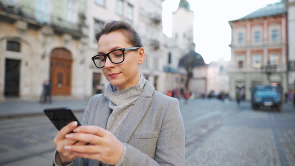 Woman with Glasses Wearing a Coat Walking Down an Old Street and Using Smartphone