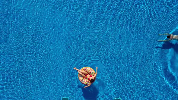 Aerial View of Man Swims in the Pool While Girl Is Lying on a Donut Pool Float