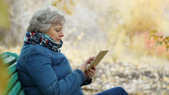 Elderly Woman Walks in the Park in Autumn Using a Digital Tablet, Online in Social Networks.