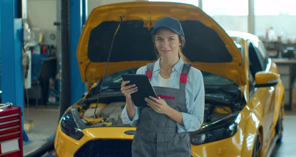 A Woman Mechanic Uses a Digital Tablet in an Auto Repair Garage. Mechanics Overhauling an Engine in