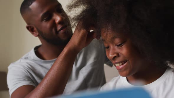 African american father tying his daughters hair while sitting on the bed at home