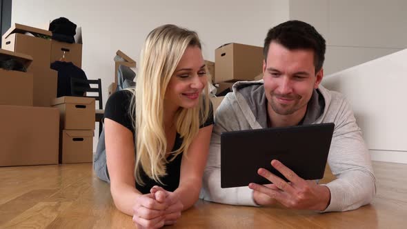 A Smiling Moving Couple Lies on the Floor of an Empty Apartment and Works on a Tablet