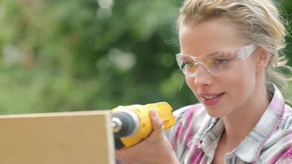 Woman wearing safety goggles drilling plywood with electric drill