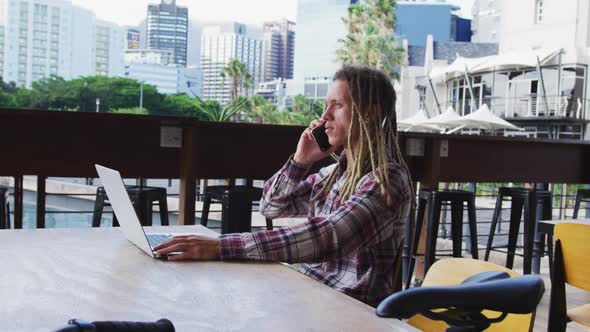 Mixed race man with dreadlocks sitting at table outside cafe using laptop and smartphone