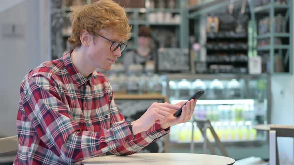 Young Redhead Man Celebrating Success on Smartphone in Cafe 