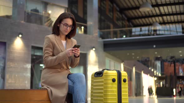 A Young Woman Sits with a Phone in Her Hands in a Shopping Center with a Suitcase. Girl in Pink Coat