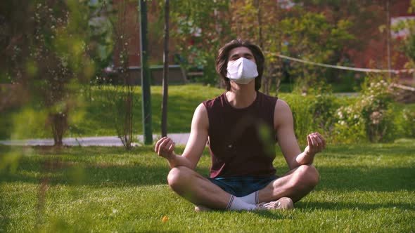 A Young Man in the Medical Mask Sitting on the Grass in the Park and Meditating