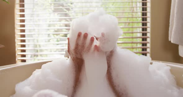 Portrait of happy biracial woman lying in bathtube, blowing foam and pampering herself