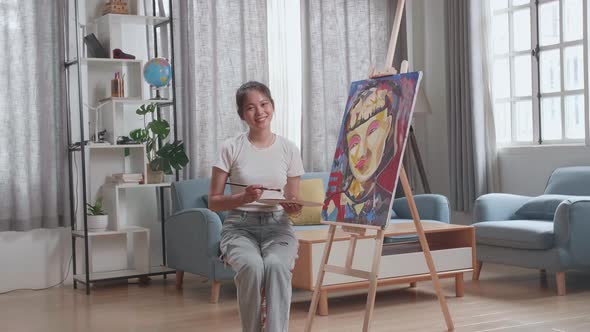 Asian Artist Girl Sitting, Holding Paintbrush And Smiling After Finish Painting A Girl On The Canvas