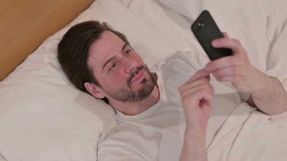 Casual Young Man Having Success on Smartphone in Bed