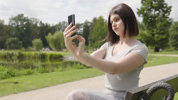 A Young Caucasian Woman Takes Selfies with a Smartphone As She Sits on a Bench in a City Park