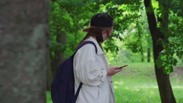 Side View of a Beautiful Woman in Cap Walking in the Park with Bagpack and Mobile Phone.