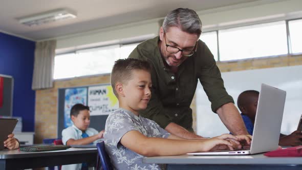 Diverse male teacher helping a schoolboy sitting in classroom using laptop