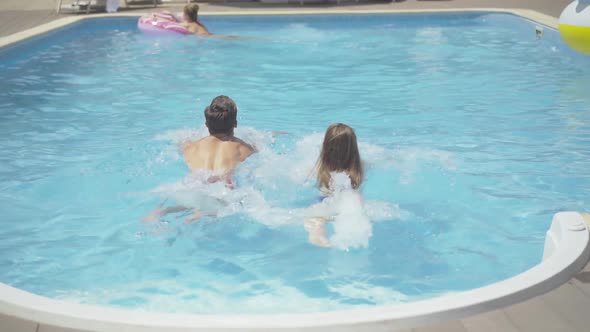 Back View of Young Caucasian Man and Woman Swimming in Outdoor Water Pool at Luxurious Resort