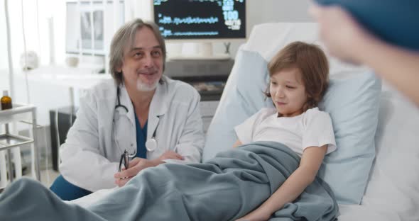 Senior Doctor Talking To Smiling Kid Patient Lying in Hospital Bed and Nurse Writing Information