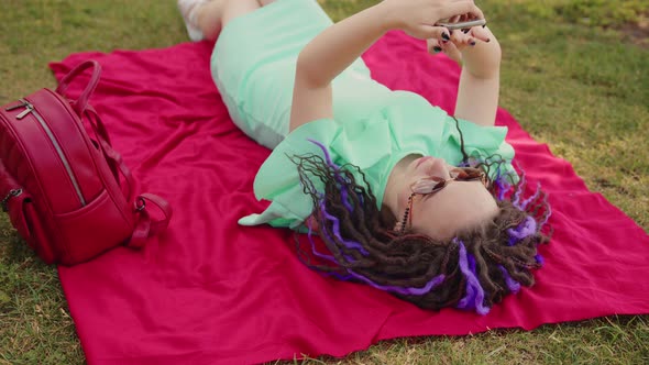 Young Woman Lying on a Blanket Blowing Uses a Smartphone in the Park