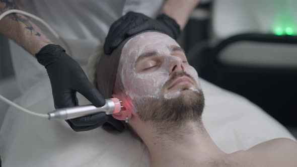 Beautician Doing Red LED Light Peel on Face of Young Handsome Man with Closed Eyes