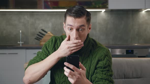 Shocked Man Reacting on Terrible Message on Smartphone Covers His Mouth in Fright Sitting on Sofa at