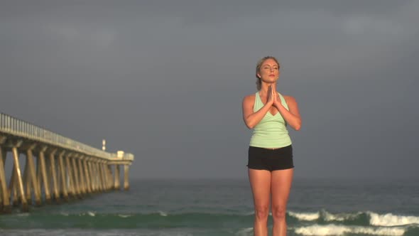 A young woman does yoga on the beach next to a pier.