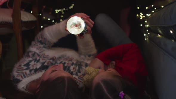 Caucasian mother and daughter holding snow globe while lying under blanket fort during christmas at