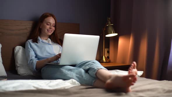 Portrait of Cheerful Red-haired Young Woman Lying on Bed and Using Laptop for Using Social Media.