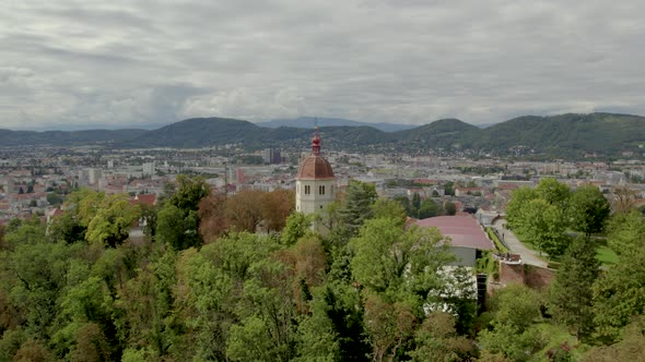 Aerial push towards Glokenturm tower on wooded hilltop with Graz's Schloßberg Austria mountain skyli